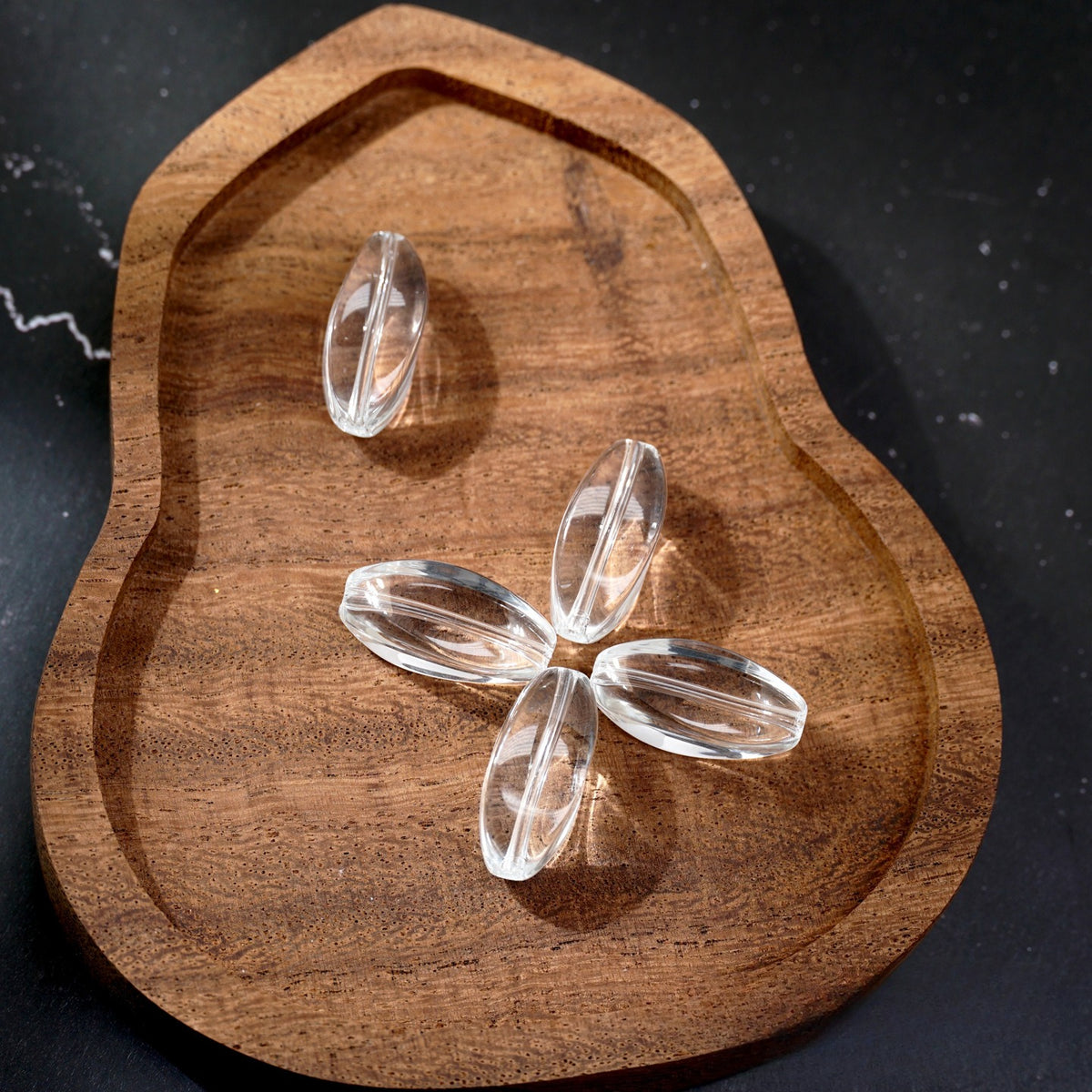Four clear teardrop-shaped glass beads on a brown wooden tray against a dark background