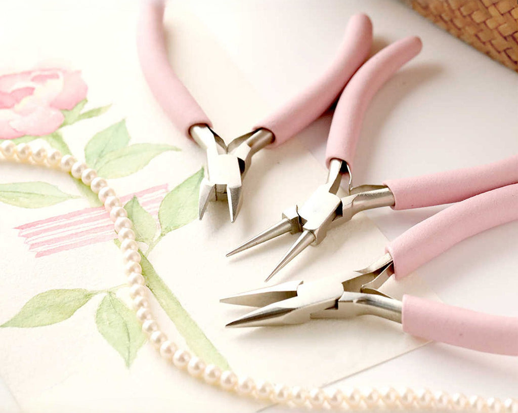 Three pink-handled jewelry pliers with silver metal tips beside pearl beads
