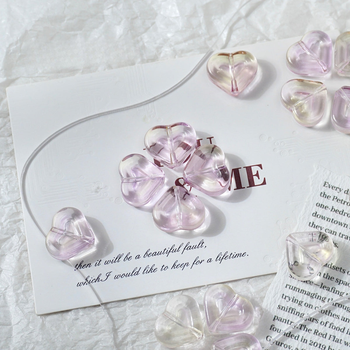 Clear heart-shaped glass beads with a soft pink tint, arranged on a white card with printed text over a textured white fabric background.