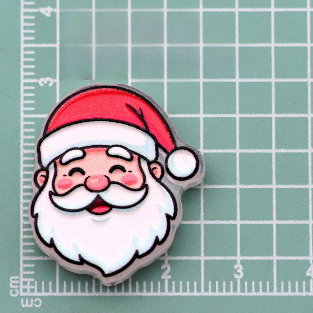 Smiling Santa face acrylic charm with red hat, white beard, and pink cheeks on green cutting mat