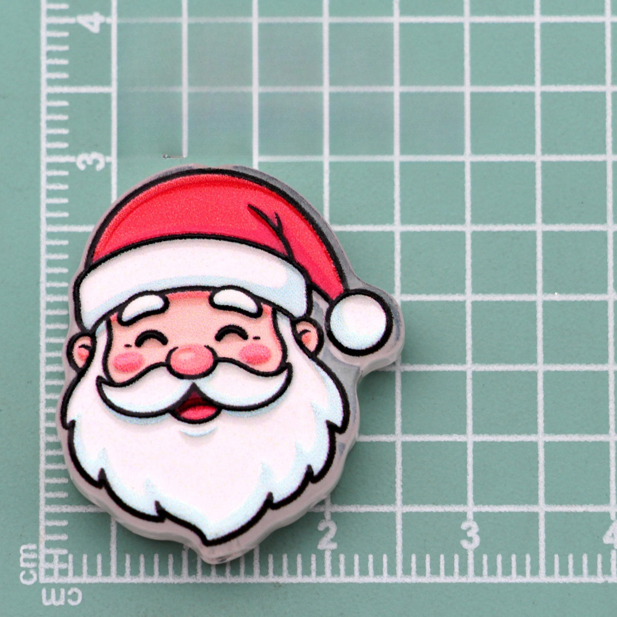 Smiling Santa face acrylic charm with red hat, white beard, and pink cheeks on green cutting mat