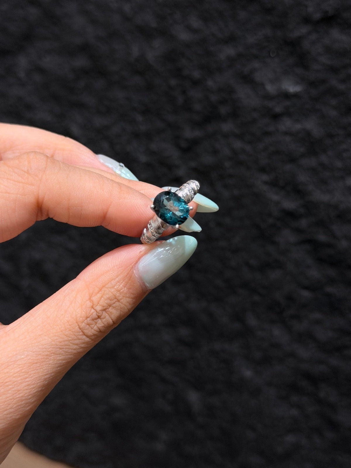 A hand with light blue manicured nails holds a silver ring featuring a round-cut deep blue gemstone against a dark textured background.