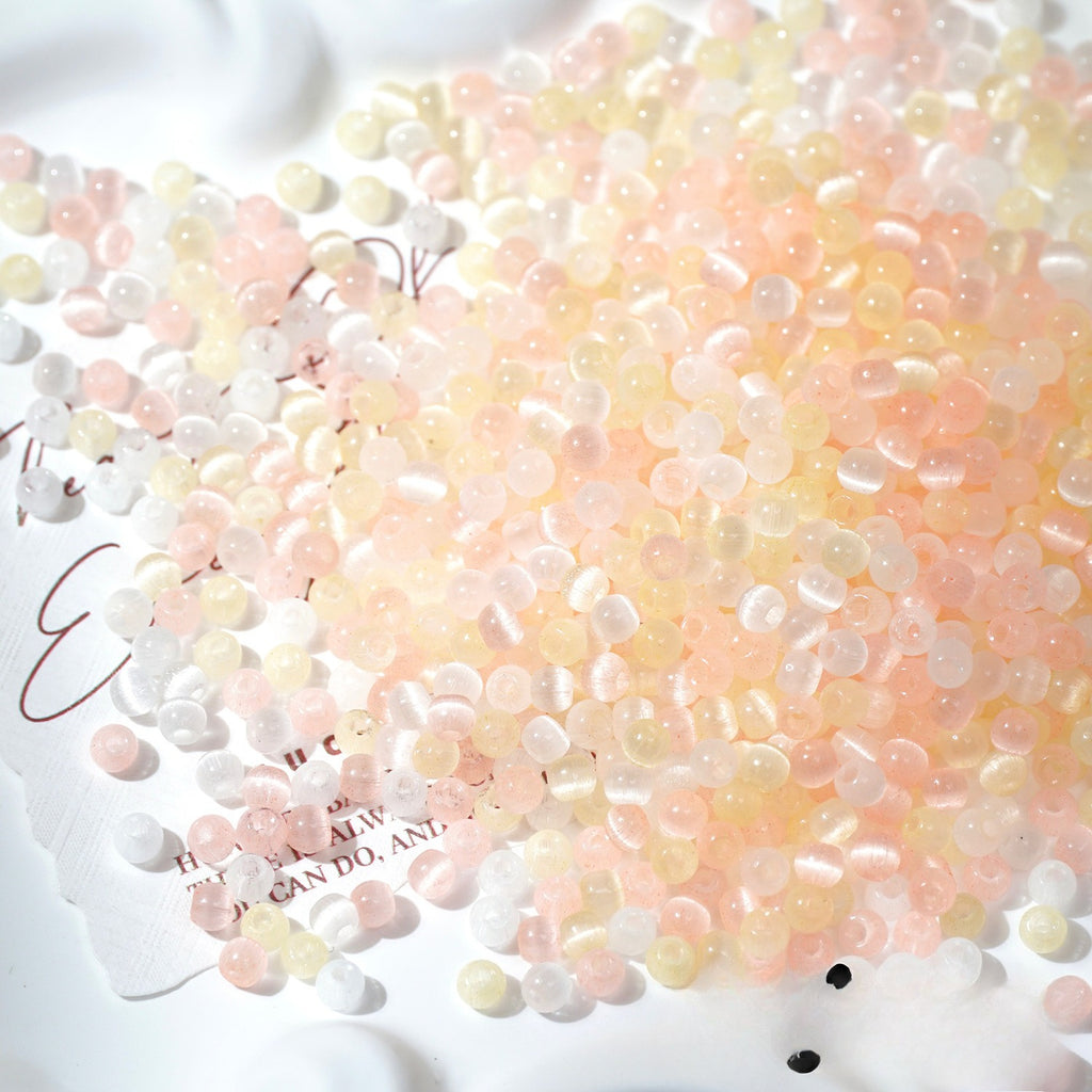 A pile of small translucent decorative beads or pebbles in soft pastel pink, peach, pale yellow, and white, scattered on a white surface with printed text partially visible underneath.