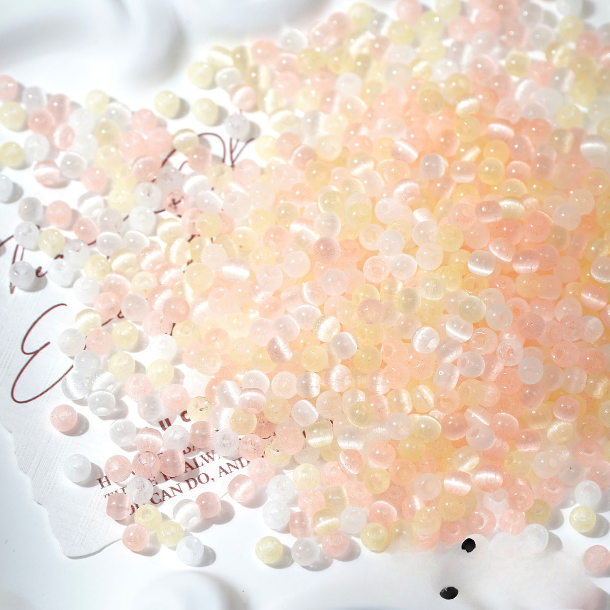 A pile of small translucent decorative beads or pebbles in soft pastel pink, peach, pale yellow, and white, scattered on a white surface with printed text partially visible underneath.