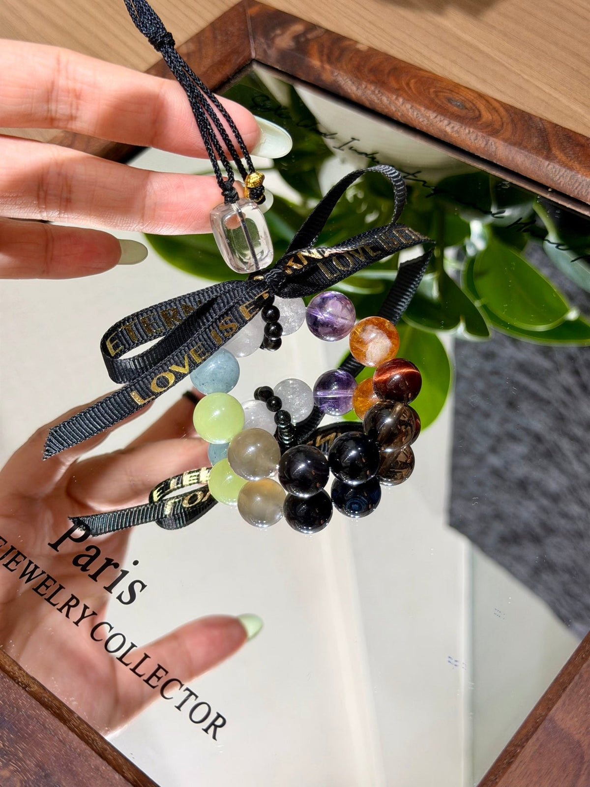 Hand holding multi-gemstone beaded car charm with black ribbon bow printed LOVE IS in gold lettering