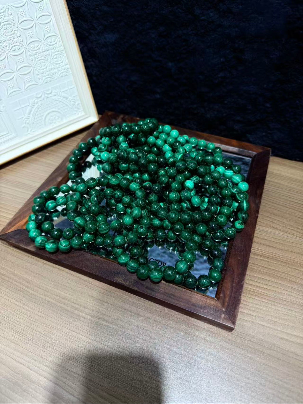 Multiple green malachite bead bracelets piled in a dark wooden tray on a light wood table, with a white decorative frame behind