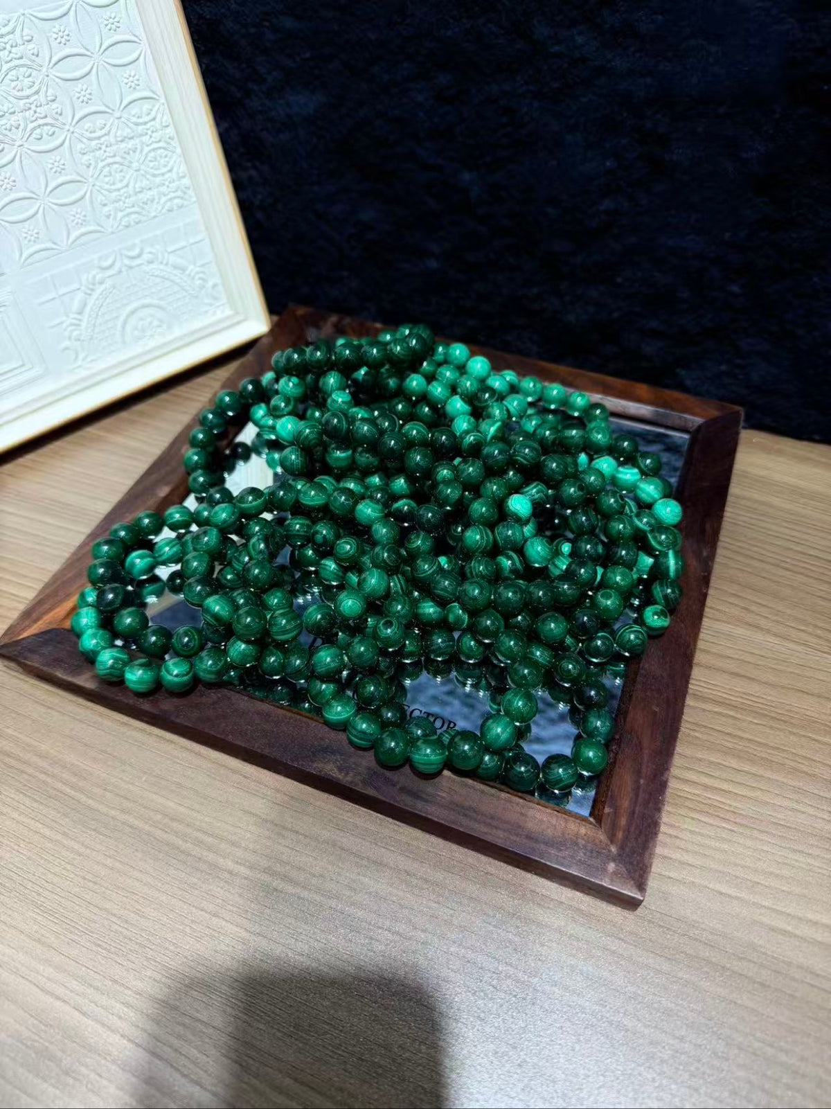 Multiple green malachite bead bracelets piled in a dark wooden tray on a light wood table, with a white decorative frame behind