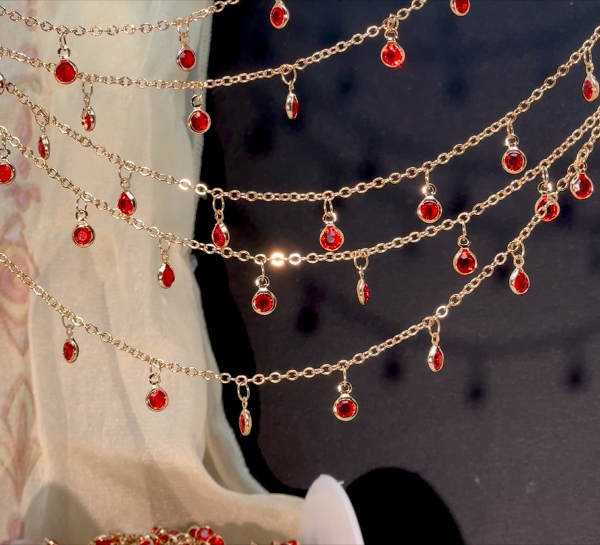 Gold-toned chain garlands with dangling red teardrop gemstones, draped across a dark background with a light fabric drape on the left.