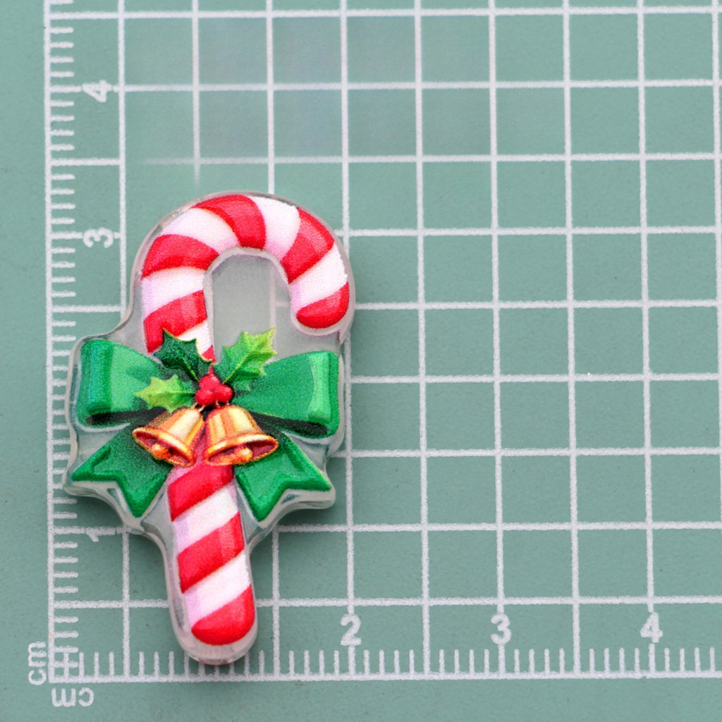 Candy cane acrylic charm with red and white stripes, green bow, holly, and gold bells on green cutting mat