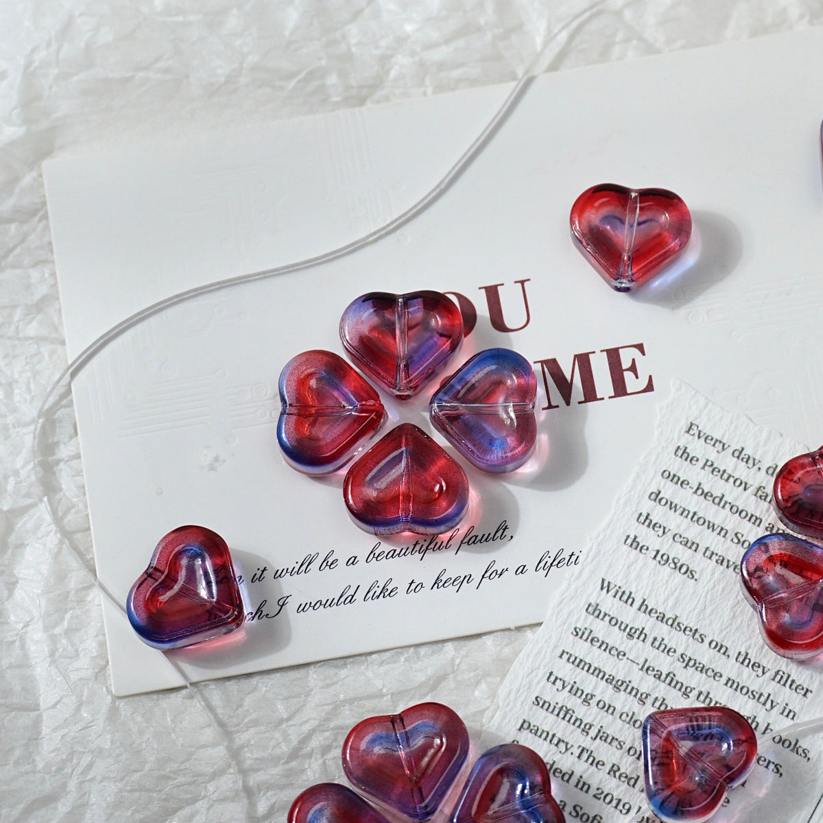 Heart-shaped translucent glass beads with a red and purple gradient swirl pattern, arranged on a white textured fabric background with printed paper cards.