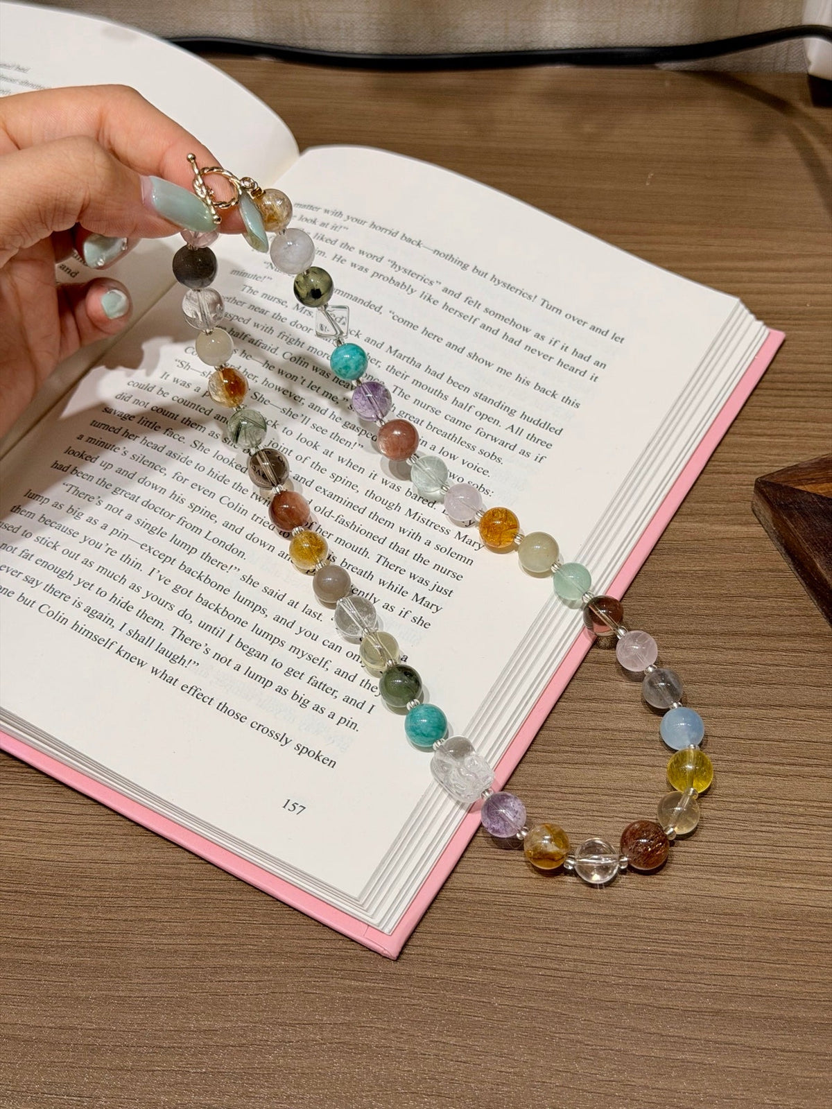 Multicolor beaded gemstone necklace draped over open book, hand holding one end, wooden tabletop