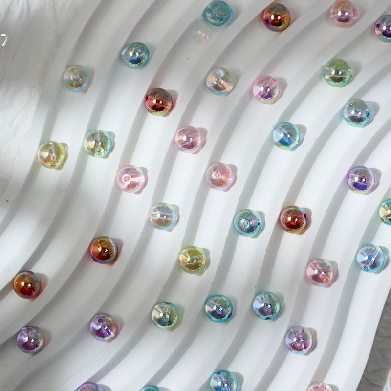 A close-up of many small iridescent oval beads or cabochons arranged on a white wavy surface. The beads reflect multiple colors including pink, blue, purple, green, and gold, with no person, animal, or real environment visible.