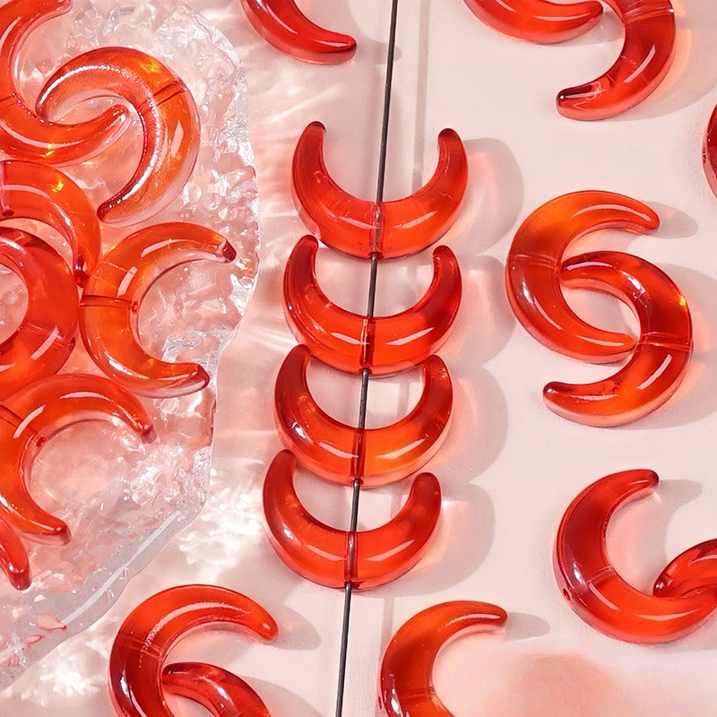 Red translucent glazed glass crescent moon beads, some strung on a wire and others on a pink surface with a clear dish