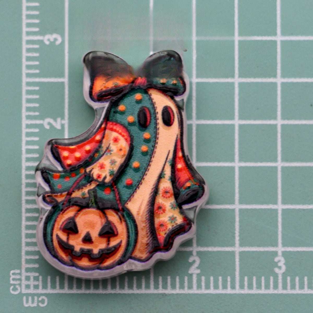 Halloween acrylic bead of a ghost with a bow holding a jack-o'-lantern pumpkin on a green cutting mat