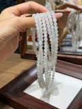 A hand holds several strands of pale blue-white beaded bracelets with a subtle iridescent sheen, draped over a wooden display tray