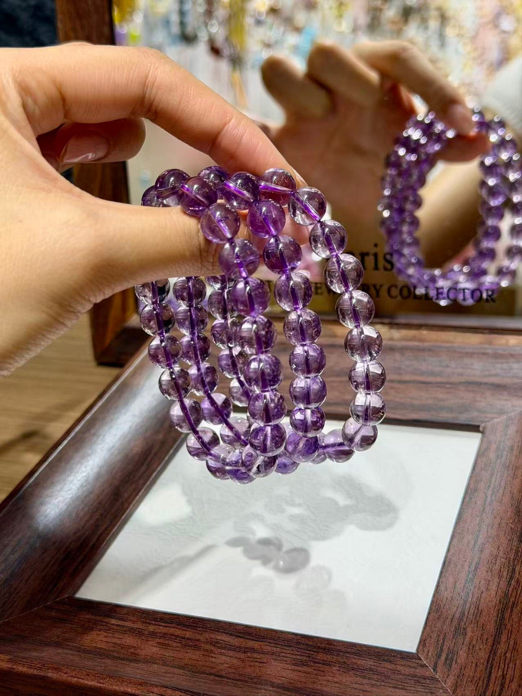 Hand holding stacked purple amethyst beaded bracelets before a wooden-framed mirror