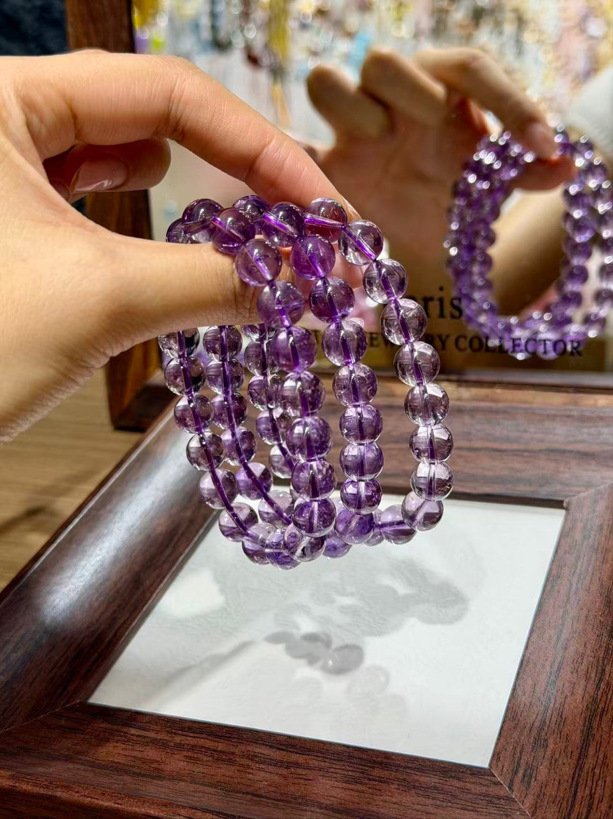 Hand holding stacked purple amethyst beaded bracelets before a wooden-framed mirror