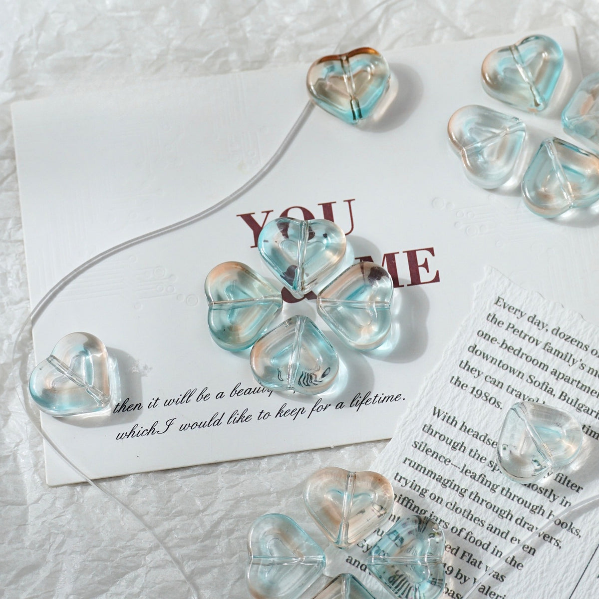 Clear heart-shaped glass beads with a light blue iridescent tint, arranged on a white textured fabric alongside printed cards.