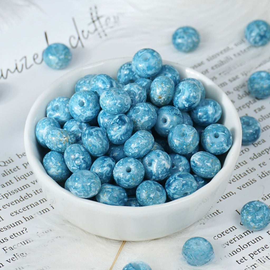 Light blue marbled round beads with drilled holes in a small white porcelain bowl, a few scattered on printed paper