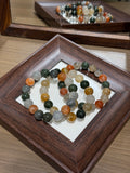 Multicolor rabbit-hair quartz bead bracelet coiled in a circle inside a dark wooden tray, with round beads in milky white, amber, gold, gray, and dark green; a mirror behind reflects the bracelet