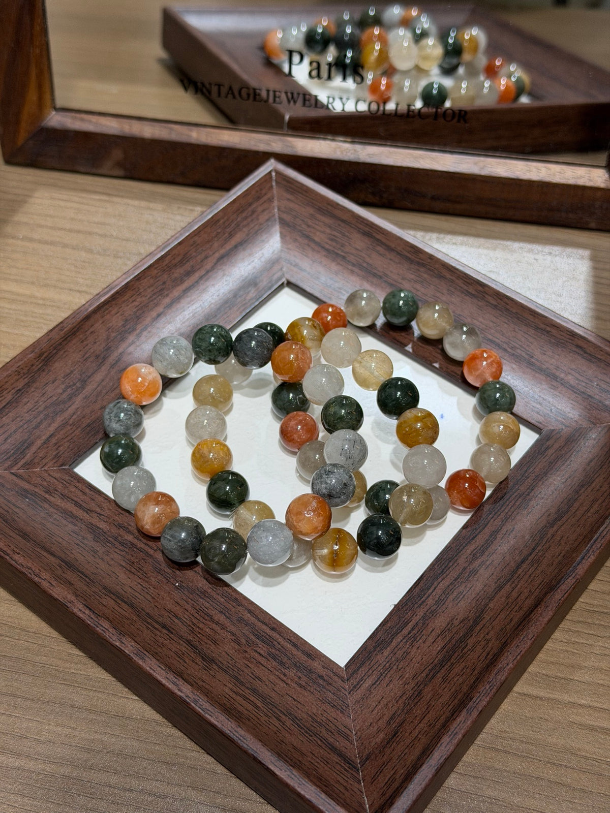 Multicolor rabbit-hair quartz bead bracelet coiled in a circle inside a dark wooden tray, with round beads in milky white, amber, gold, gray, and dark green; a mirror behind reflects the bracelet
