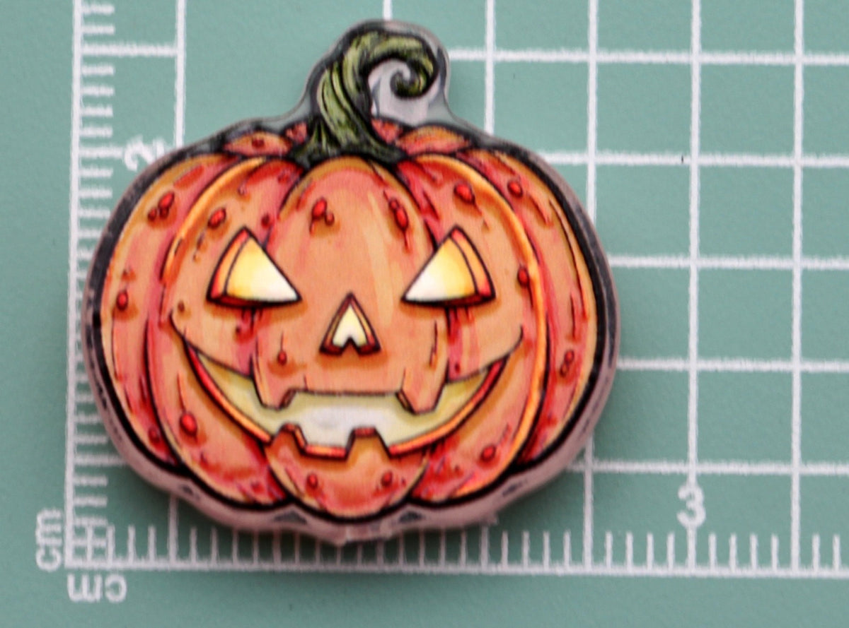 Halloween pumpkin-shaped acrylic charm with carved jack-o'-lantern face on green cutting mat with centimeter grid