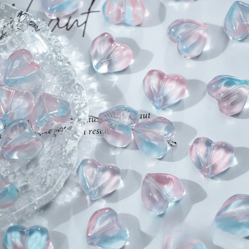 Heart-shaped translucent glass beads with a pink and blue ombré finish, scattered on a white surface with a decorative clear glass dish on the left.