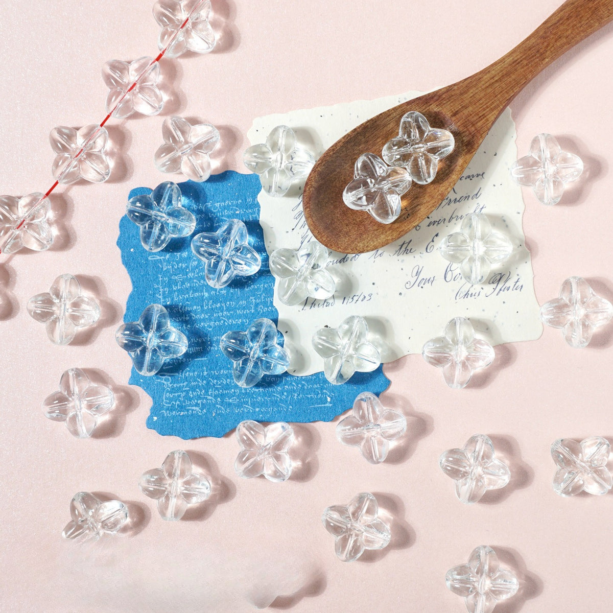 Clear four-petal flower glass beads on light pink with a wooden spoon and blue and white paper